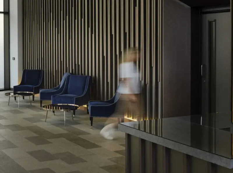 An office waiting room: three blue armchairs, wood wall, person walking by.