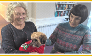 Two women smile to camera standing up in room. The woman on the left is holding a dog which is wearing a red Christmas Jumper with a tree on. The other woman is wearing a Christmas jumper and petting the dog.